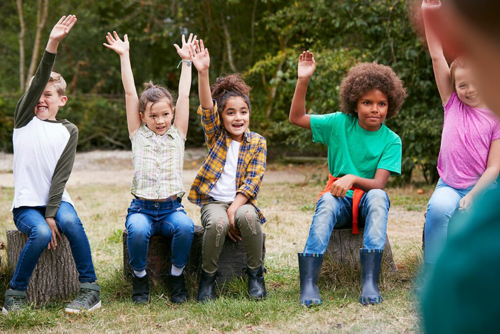 Eine Gruppe Kinder sitzen auf Baumstümpfen und halten die Hände hoch