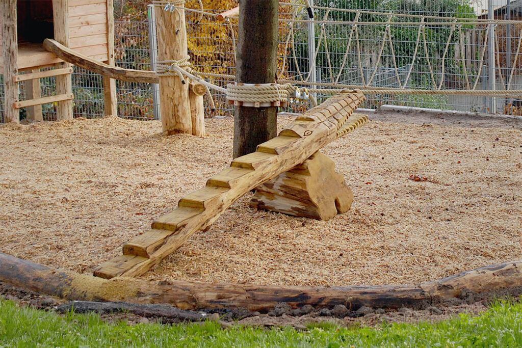 Ein hölzernes Spielplatzgerüst mit einer Krokodilaufstiegsrampe, Baumstämmen und Seilen steht auf einem Bett aus Holzspänen. Das Gelände ist von einem Drahtzaun umgeben, mit Gras im Vordergrund und einem Holzschuppen auf der linken Seite des Bildes.