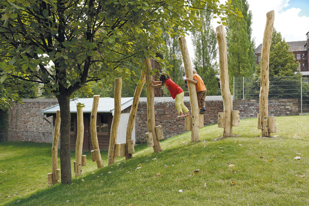 Zwei Kinder spielen am Stelzenweg und balancieren und klettern auf senkrechten Holzpfosten, die entlang eines Grashügels aufgereiht sind. Grüne Bäume, eine Backsteinmauer und ein Gebäude mit Zaun schaffen eine lebendige Szene unter dem teilweise bewölkten Himmel.