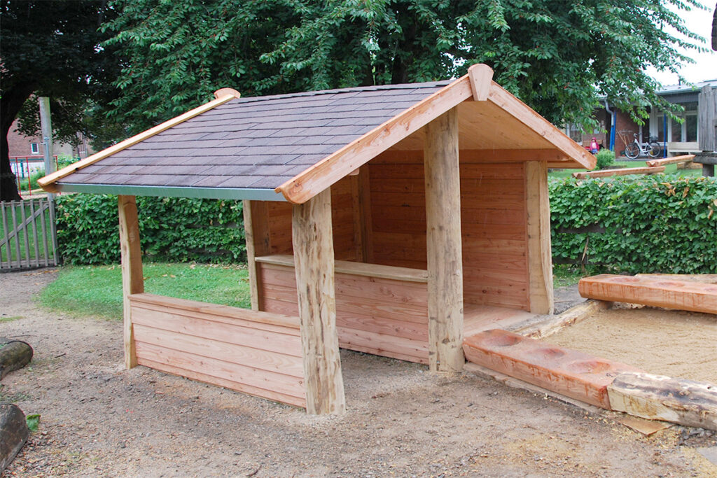 Ein hölzernes Spielhaus mit schrägem Dach steht wie ein gemütlicher Drive-in-Platz neben einem Sandkasten auf einem Spielplatz im Freien. Es ist an zwei Seiten offen und aus Naturstämmen und Brettern gebaut, eingebettet zwischen Bäumen, einer Hecke und einem Zaun.