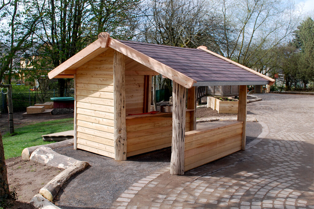 Ein kleines hölzernes Spielhaus mit offener Front und Seite, gebaut aus Holzstämmen und Brettern, steht auf einem geschwungenen Kopfsteinpflasterweg wie ein kleines Autokino. Bäume und Gras umgeben den Bereich, mit Holzbänken und Pflanzgefäßen in der Nähe in einer parkähnlichen Umgebung.