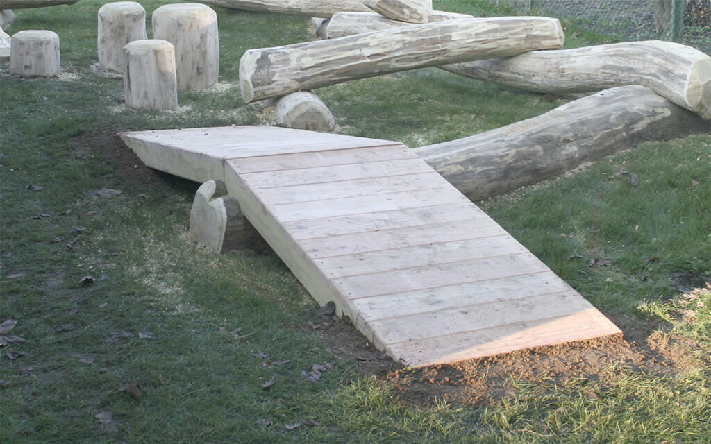 Eine Holzrampe, die Rampe am Baumstamm zweiseitig gestaltet, führt zu großen, glatten Baumstammstrukturen auf grasbewachsenem Boden. Im Hintergrund vervollständigen aufrechte Baumstammhocker diese natürliche Spielplatz- oder Hindernisbahnanlage.