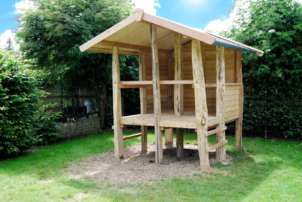 Ein Holzspielhaus von Pfahlhaus Klein mit schrägem Dach steht auf Stelzen in einem grasbewachsenen Garten, umgeben von grünen Büschen. Die nach vorne offene Konstruktion hat teilweise geschlossene Seiten, eine kleine Leiter und Sonnenlicht, das spielerische Schatten wirft.