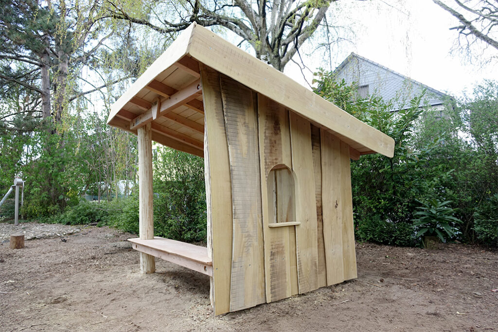 Ein kleines hölzernes Handwerkerhaus mit steilem, schrägem Dach steht auf kahlem Boden. Es verfügt über eine Bank und ein Bogenfenster an einer Wand, umgeben von Bäumen, Büschen und einem Nachbarhaus im Hintergrund.