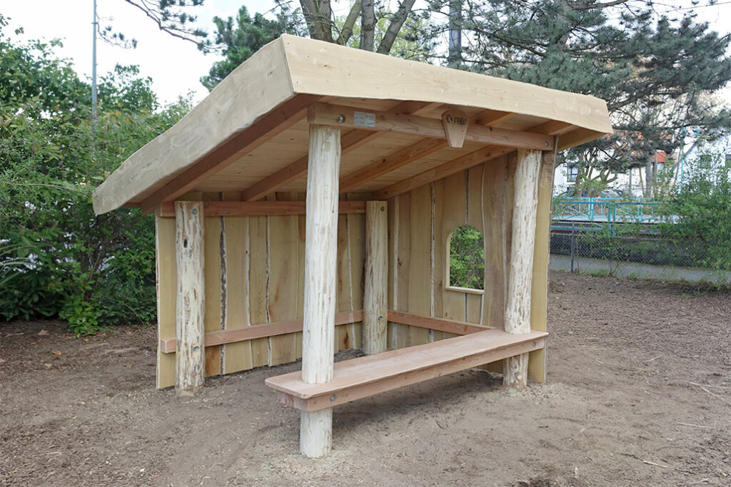 Ein kleines hölzernes Handwerkerhaus mit schrägem Dach, das von Rundholzpfeilern gestützt wird und an zwei Seiten über eingebaute Bänke verfügt. Das Gebäude hat Teilwände, einen quadratischen Fensterausschnitt und steht auf nacktem Boden, umgeben von Bäumen und Grünpflanzen.