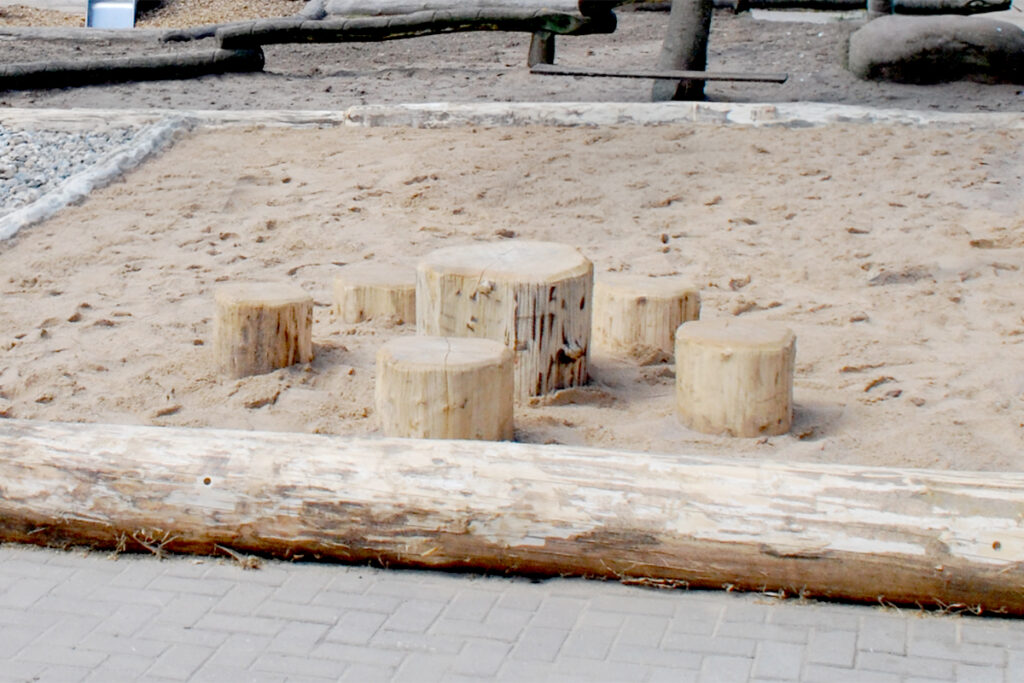 Auf einem sandigen Spielplatz befindet sich eine Stammholzsitzgruppe - Baumstümpfe in verschiedenen Höhen, die wie Sitzgelegenheiten angeordnet sind. Glatte Baumstämme begrenzen den Sandkasten, im Vordergrund ist ein gepflasterter Weg und im Hintergrund sind hölzerne Spielgeräte zu sehen.